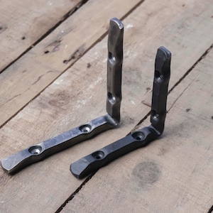 May include: Two black metal shelf brackets with a rustic finish. The brackets are L-shaped and have four holes on each side for mounting.