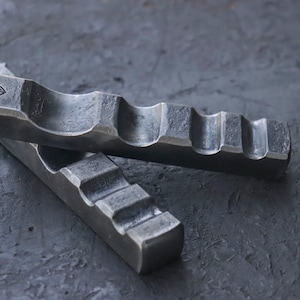 May include: Two metal tools with a textured surface and a series of grooves. The tools are made of a dark gray metal and are lying on a dark gray surface.