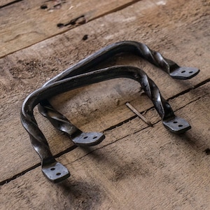 May include: Two black metal twisted handles with two holes on each end. The handles are on a wooden surface.