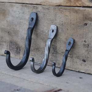 May include: Three black metal hooks with a twisted design. The hooks are different sizes and are mounted on a wooden surface.