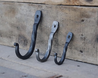 May include: Three black metal hooks with a twisted design. The hooks are different sizes and are mounted on a wooden surface.