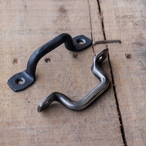 May include: Two metal handles, one black and one silver, rest on a weathered wooden surface. The black handle has a textured, hammered appearance, while the silver handle has a wavy design. Both have mounting holes.