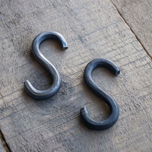 May include: Two black metal S-shaped hooks on a wooden surface.