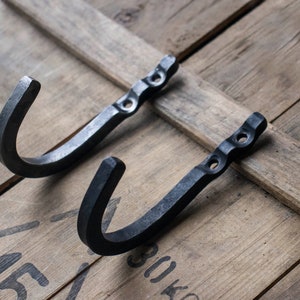 May include: Two black, hand-forged metal hooks with a curved design. Each hook has a flat, rectangular base with two holes for mounting. The hooks are displayed on a rustic, wooden surface.