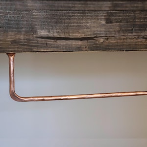 May include: A copper-colored, handcrafted toilet paper holder. The holder is mounted beneath a dark, weathered wooden shelf. The copper bar extends horizontally with a curved end to hold the paper roll. The design is rustic and industrial.