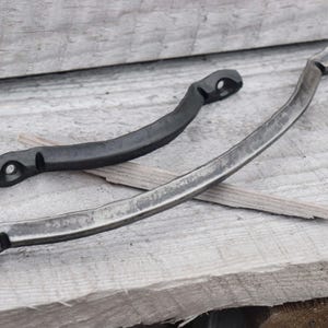 May include: Two metal handles with curved designs. One is black, the other is silver. Both have holes at each end. They rest on a weathered wooden surface, suggesting a rustic or industrial aesthetic. The handles appear to be handmade.