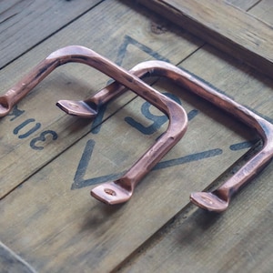 May include: Two copper-colored metal handles with a curved design. The handles are placed on a wooden surface with the number "30" stenciled in black. The handles have a rustic, industrial aesthetic.