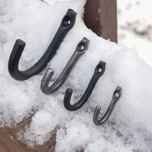 May include: Four black metal hooks of varying sizes are arranged in a row on a snowy surface. The hooks are all made of the same material and have a simple, functional design.