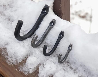 May include: Four black metal hooks of varying sizes are arranged in a row on a snowy surface. The hooks are all made of the same material and have a simple, functional design.