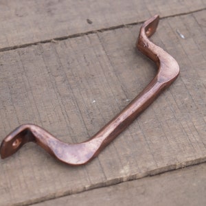 May include: A copper-colored metal door handle with a curved design. The handle is made of a textured metal and has a rustic look.