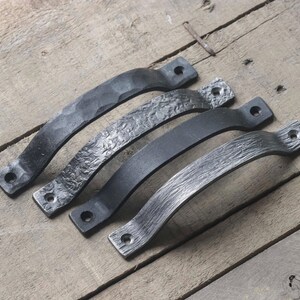 May include: Four black metal handles with varying textures, including hammered and textured finishes. Each handle has two holes for mounting. The handles are arranged diagonally on a weathered wooden surface.