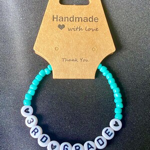 May include: A turquoise beaded bracelet with white beads spelling "3rd Grade" with hearts on either side. The bracelet is on a brown card that says "Handmade with love" and "Thank You".