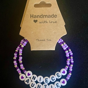 Könnte beinhalten: Zwei Armbänder mit Perlen, die die Wörter "NIECE" und "AUNTIE" mit weißen Buchstabenperlen schreiben. Die Armbänder sind lila und silberfarben, mit einem Herzsymbol. Sie sind an einem braunen Anhänger mit dem Text "Handmade with love" und "Thank You" befestigt.