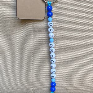 Puede incluir: Un llavero de cuentas con las palabras "DUE PROCESS" escritas con cuentas de letras blancas. Las cuentas están ensartadas en un cordón azul e intercaladas con cuentas azules de diferentes tonos. El llavero está sujeto a una bolsa beige.