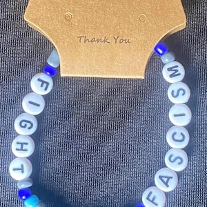 May include: A handmade bracelet with white letter beads spelling "FIGHT FASCISM". The bracelet features blue, gray, and turquoise beads. A brown tag above the bracelet reads "Handmade with love" and "Thank You".