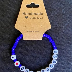 May include: A blue beaded bracelet with white letter beads spelling "I DUE PROCESS" and a red heart bead. The bracelet is displayed with a brown tag that reads "Handmade with love" and "Thank You".