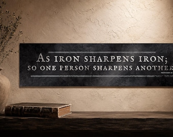 May include: A rectangular black wall sign with a textured finish featuring the white text: As iron sharpens iron; so one person sharpens another. Proverbs 27:17. The sign includes thin horizontal white lines above and below the main text.