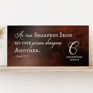 Könnte beinhalten: Ein rustikales Schild aus braunem Metall mit einer weißen Inschrift, die lautet: "Wie Eisen Eisen schärft, so schärft eine Person die andere. Sprichwörter 27:17". Das Schild zeigt auch einen weißen kursiv geschriebenen Buchstaben "C" und den Text "Lacey & Eric Crane 03.28.2003".