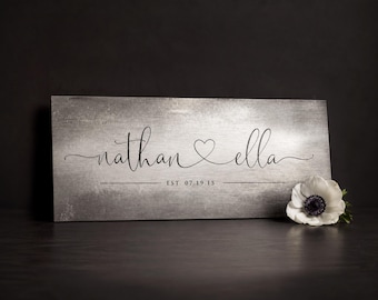 May include: A rectangular gray sign with a brushed metal texture featuring the black script text nathan and ella separated by a heart outline. Below the names, the text EST. 07.19.15 is centered between two horizontal lines. A white flower is attached.