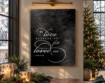 May include: A rectangular wall art piece featuring a dark, textured black background with white cursive and block lettering that reads: We love because He first loved us 1 John 4:19. The text is centered on the canvas.