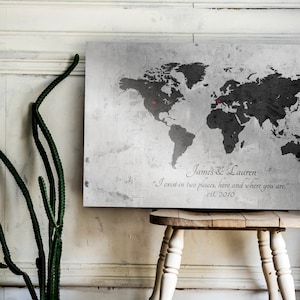 May include: A gray world map print with two red push pins marking locations. The print has a distressed, vintage look and includes the text "James & Lauren" and "I exist in two places, here and where you are." with the date "est. 2010".