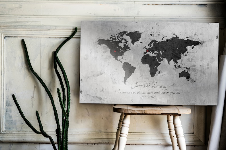 May include: A gray world map print with two red push pins marking locations. The print has a distressed, vintage look and includes the text "James & Lauren" and "I exist in two places, here and where you are." with the date "est. 2010".