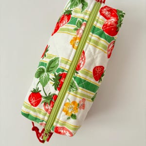 Peut inclure: Trousse rectangulaire avec un motif de fraises matelassé. La trousse présente des fraises rouges, des feuilles vertes et des fleurs jaunes sur un fond blanc avec des rayures vertes. Elle a une fermeture éclair verte et une tirette à carreaux rouges et blancs.