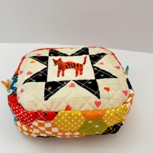 May include: A quilted, square pouch with a patchwork design. The top has a star pattern with an orange tiger image. The sides are a mix of colourful fabrics, including polka dots and floral prints. A small loop is attached.