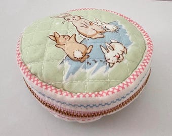 Bolsita de tela acolchada con cremallera, hecha a mano, con estampado de conejitos y forma redonda pequeña.