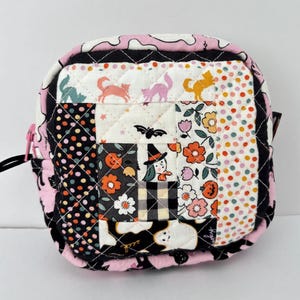 May include: A small, square, quilted pouch with a black zipper and pull. The patchwork design features Halloween-themed patterns, including cats, bats, pumpkins, and a witch. The edges are trimmed with pink fabric.