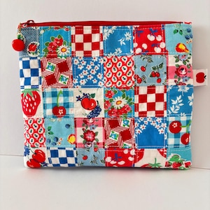 Pode incluir: Uma bolsa retangular com fecho de correr apresentando um design de patchwork de vários tecidos de algodão em vermelho, branco e azul. Os padrões incluem morangos, cerejas, flores e quadrados. Um pequeno pendente de cereja está no fecho.