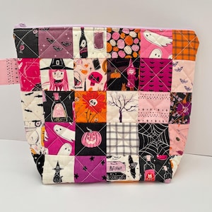 Peut inclure: Une pochette zippée matelassée avec un motif patchwork de carrés de tissu sur le thème d'Halloween. Le design présente des illustrations de fantômes, citrouilles, sorcières, chauves-souris, toiles d'araignée, potions et arbres en rose, orange, noir et violet.