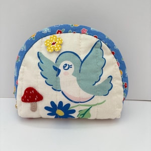 May include: A semi-circular quilted fabric pouch featuring a light blue bird, a red mushroom, and a blue flower on an off-white background. The pouch is bordered by a blue fabric trim with a floral pattern and includes a yellow flower-shaped button.