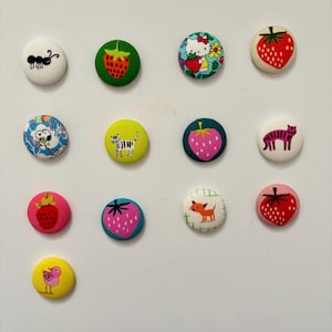 Peut inclure: Une collection de douze boutons recouverts de tissu avec divers motifs. Les boutons présentent des images de fraises, d'une fourmi, d'un tigre, d'un chien, d'un oiseau et d'un personnage de dessin animé. Les boutons sont de différentes couleurs, dont le rouge, le vert, le rose, le jaune et le bleu.