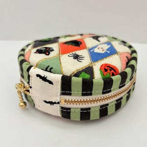 Può includere: Una pochette rotonda in tessuto trapuntato con cerniera, caratterizzata da un motivo a rombi con icone di Halloween tra cui fantasmi, zucche, ragni e gatti neri. Il pannello laterale presenta strisce nere e verdi e una linguetta bianca.