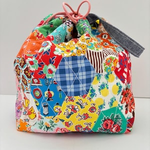 Könnte beinhalten: Ein Kordelzugbeutel aus gestepptem Stoff mit einem Patchwork aus sechseckigen Feldern in verschiedenen Farben und Mustern, darunter Blumen-, Karomuster und Küchenmotive. Die Tasche hat einen rosa Kordelzugverschluss und einen dunkelblauen Jeanshenkel.
