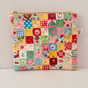 Puede incluir: Un estuche cuadrado acolchado con cremallera que presenta un diseño de patchwork de varios cuadrados de tela coloridos. Los patrones incluyen fresas, cerezas, gatitos, lazos y cuadros en tonos rojo, amarillo, azul, verde y rosa. Una pequeña etiqueta blanca dice hand made.