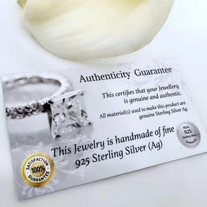 May include: A white card with the text "Authenticity Guarantee" and "This certifies that your Jewellery is genuine and authentic. All material(s) used to make this product are genuine Sterling Silver Ag. This Jewelry is handmade of fine 925 Sterling Silver (Ag)". A silver ring with a square-shaped stone is pictured on the card. The card also has a 100% Satisfaction Guarantee seal.