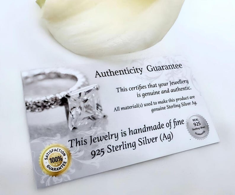 May include: A white card with the text "Authenticity Guarantee" and "This certifies that your Jewellery is genuine and authentic. All material(s) used to make this product are genuine Sterling Silver Ag. This Jewelry is handmade of fine 925 Sterling Silver (Ag)". A silver ring with a square-shaped stone is pictured on the card. The card also has a 100% Satisfaction Guarantee seal.