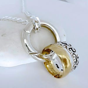 May include: A silver chain necklace featuring a pendant composed of three interlocking rings. One ring is polished silver, one is gold-toned with a smooth finish, and the third is silver with an intricate, repeating black-filled scrollwork pattern.