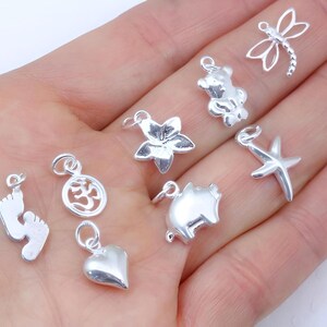 May include: A collection of eight small silver-toned metal charms featuring various shapes including a dragonfly, a teddy bear, a flower, a starfish, a pig, a heart, an Om symbol, and a pair of baby feet, each with a small circular jump ring.