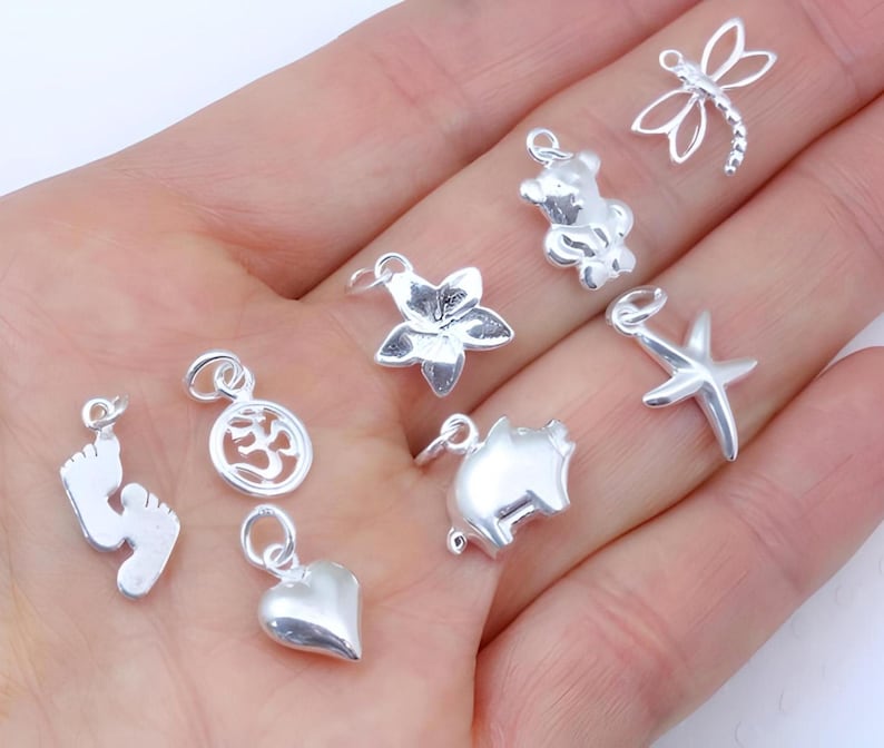 May include: A collection of eight small silver-toned metal charms featuring various shapes including a dragonfly, a teddy bear, a flower, a starfish, a pig, a heart, an Om symbol, and a pair of baby feet, each with a small circular jump ring.