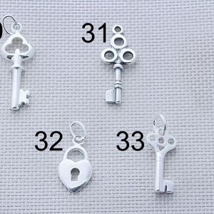 Puede incluir: Cuatro dijes de metal plateado numerados 30, 31, 32 y 33. El dije 30 es una llave antigua, el 31 es una llave decorativa con cabeza circular, el 32 es un candado en forma de coraz&oacute;n y el 33 es una llave con una cabeza circular calada y adornada.