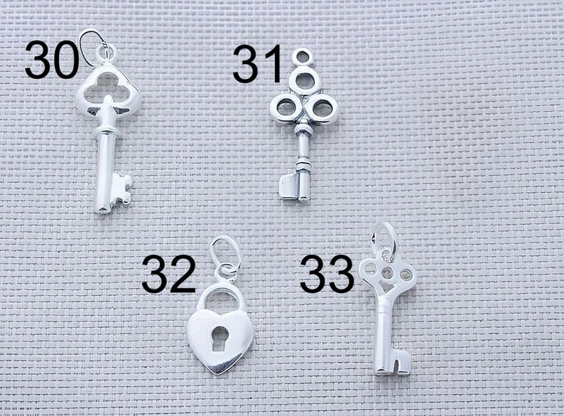 Puede incluir: Cuatro dijes de metal plateado numerados 30, 31, 32 y 33. El dije 30 es una llave antigua, el 31 es una llave decorativa con cabeza circular, el 32 es un candado en forma de coraz&oacute;n y el 33 es una llave con una cabeza circular calada y adornada.