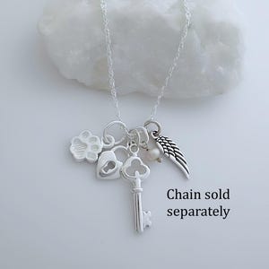 Puede incluir: Un collar de tono plateado con varios dijes que incluyen una huella de pata, un candado en forma de coraz&oacute;n, una llave antigua, una peque&ntilde;a perla blanca y un ala de &aacute;ngel. El texto "Chain sold separately" aparece en la parte inferior derecha.
