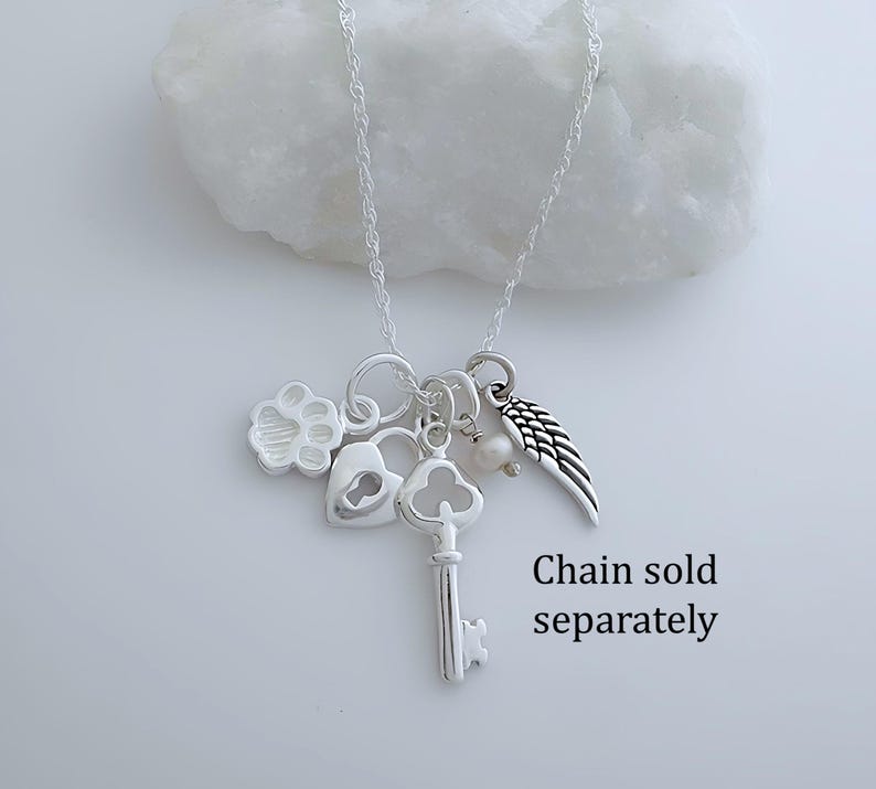 Puede incluir: Un collar de tono plateado con varios dijes que incluyen una huella de pata, un candado en forma de coraz&oacute;n, una llave antigua, una peque&ntilde;a perla blanca y un ala de &aacute;ngel. El texto "Chain sold separately" aparece en la parte inferior derecha.