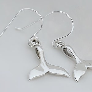 May include: A pair of polished silver-toned earrings featuring a whale tail charm design. Each earring hangs from a curved ear wire hook, showcasing a smooth, reflective metallic finish that captures light across the curved fluke surfaces.