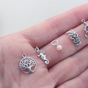 Puede incluir: Una colecci&oacute;n de cinco peque&ntilde;os dijes de metal que incluyen un &aacute;rbol de la vida circular, un caballito de mar, una perla blanca en un anillo plateado, un b&uacute;ho y un ancla de barco con tim&oacute;n, todos con un acabado plateado pulido.