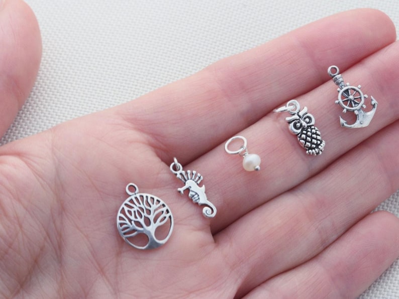Puede incluir: Una colecci&oacute;n de cinco peque&ntilde;os dijes de metal que incluyen un &aacute;rbol de la vida circular, un caballito de mar, una perla blanca en un anillo plateado, un b&uacute;ho y un ancla de barco con tim&oacute;n, todos con un acabado plateado pulido.