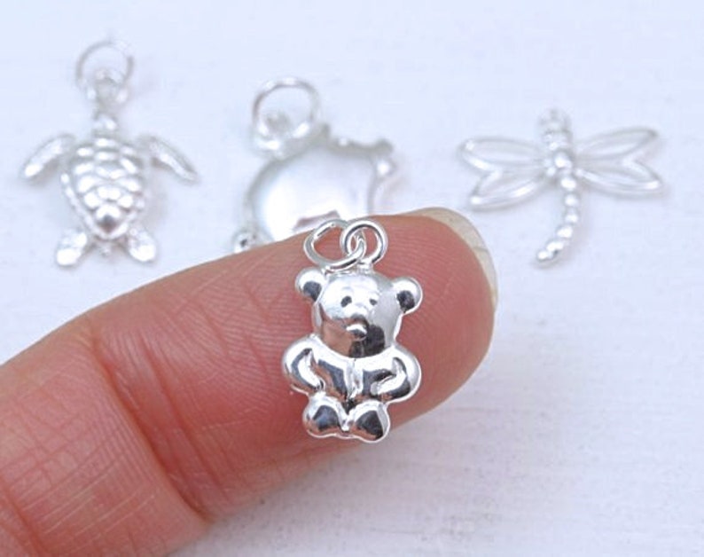 May include: A collection of small silver-toned metal charms featuring a teddy bear, a sea turtle, and a dragonfly. Each charm includes a circular jump ring at the top for attachment. The teddy bear charm is shown in the foreground with a polished finish.